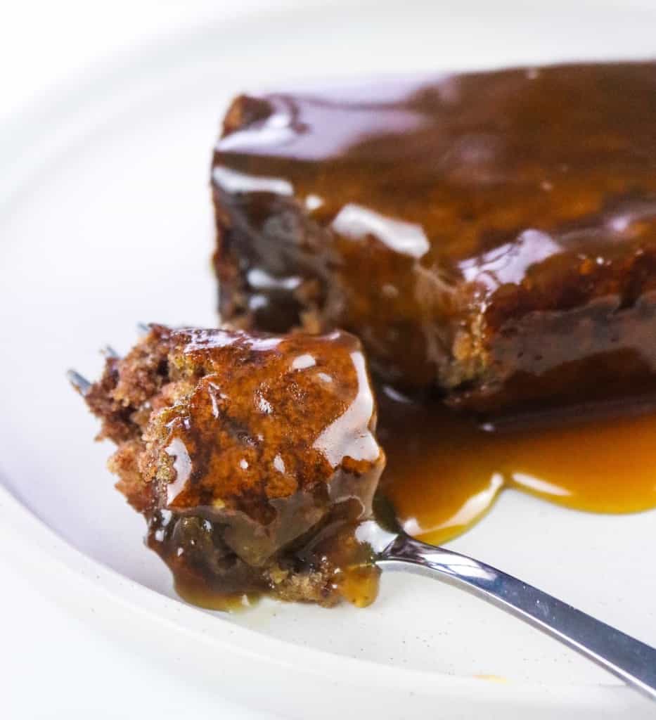 vegan sticky toffee pudding slice on a white plate with fork and oozing toffee sauce