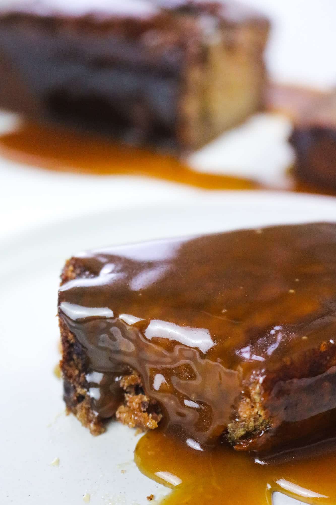 the best vegan sticky toffee pudding slice on a white plate covered in toffee sauce on focus and rest of loaf on the background