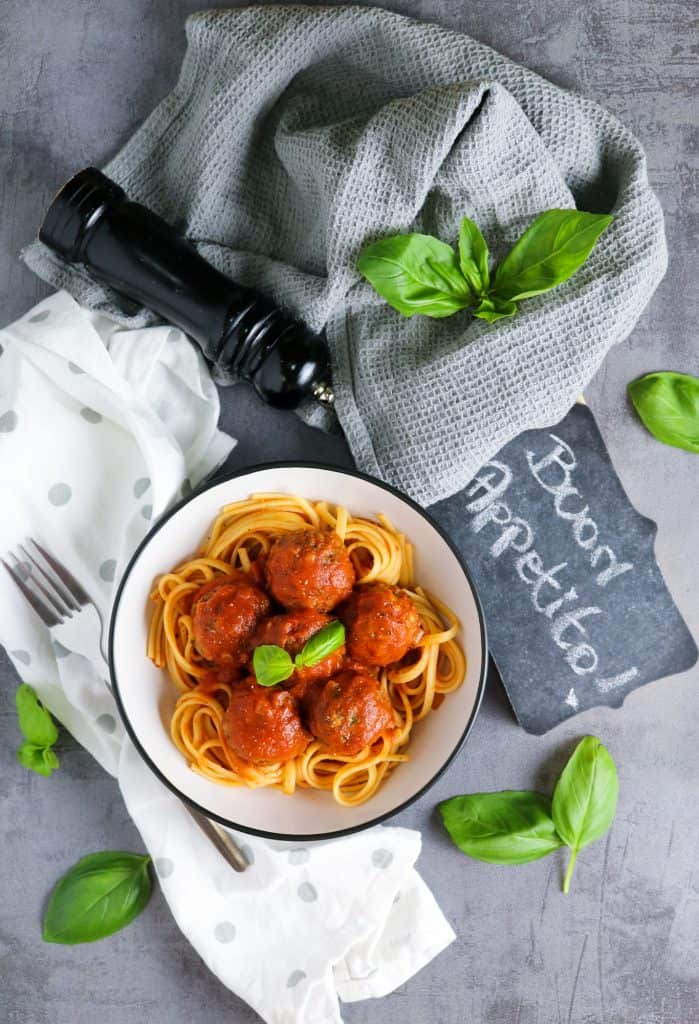 Lentil Meatballs in Rich Tomato Sauce over cooked spaghetti on a grey background with cloths, pepper grinder, "buon appetito" sign and basil leaves