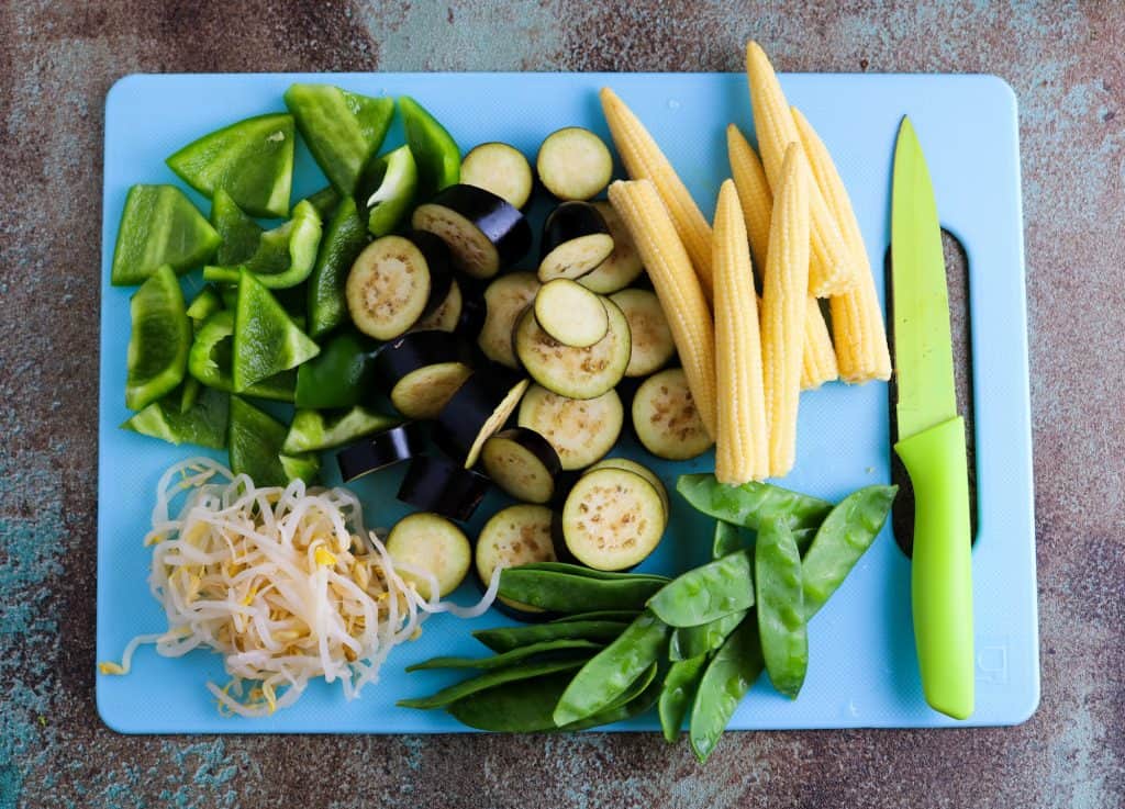 green pepper, soy, aubergine, baby corn and mangetout chopped up on a chopping board to make vegan Thai Green Curry and rice noodles