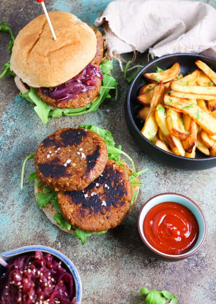 Vegan burgers patties made of soy TVP and gluten with oven baked chips on a side, ketchup and a burger on the background oozing red onion chutney