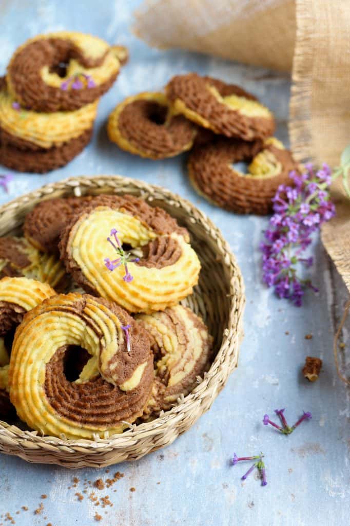 Vegan Whipped Cookies rings with cocoa powder nicely arranged on a wicker plate decorated with puple flowers bottom left