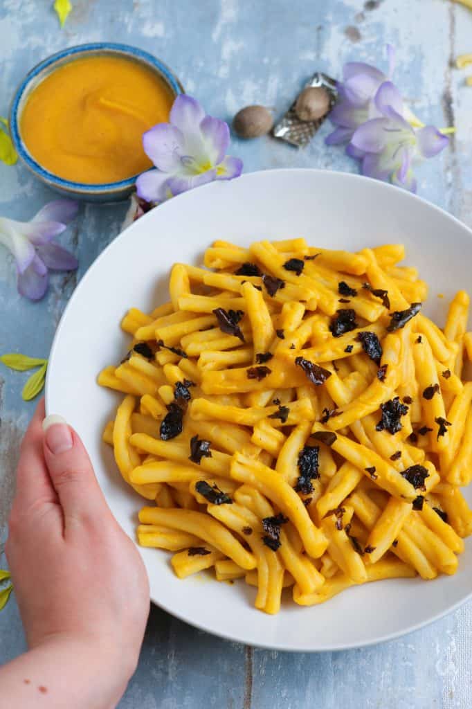 crispy dulse seaweed on pasta in a butternut squash sauce, displayed with purple and yellow flowers on a white plate and a small dish of butternut squash sauce for Butternut Squash Pasta with Crispy Smoked Seaweed