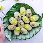 boiled potatoes with vegan tuna and egg yolk filling on a leaf shaped green plate, left hand holding the plate. All on a white background, peppercorns, lettuce and yellow flowers as decorations