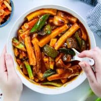 Korean Tteokbokki (떡볶이) with Peppers in a white bowl with white chinese spoon in the bowl, a bowl of kimchi in the corner, left hand holding the bowl and right hand stirring the dish with a chinese spoon