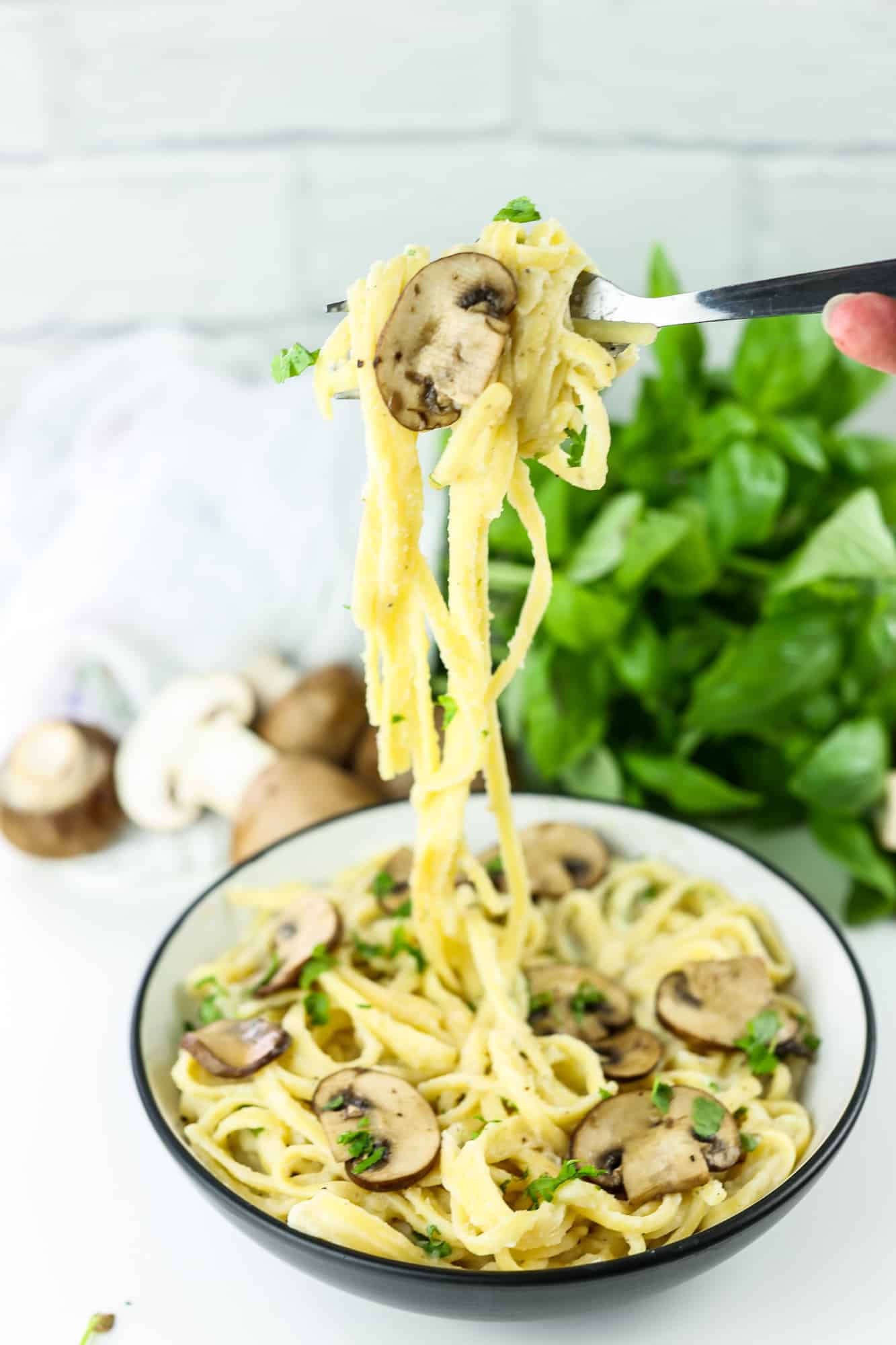 Vegan Creamy Cauliflower and Mushroom Pasta in a white bowl, right hand with a fork lifting spaghetti up above the serving bowl with raw mushrooms and basil on the background