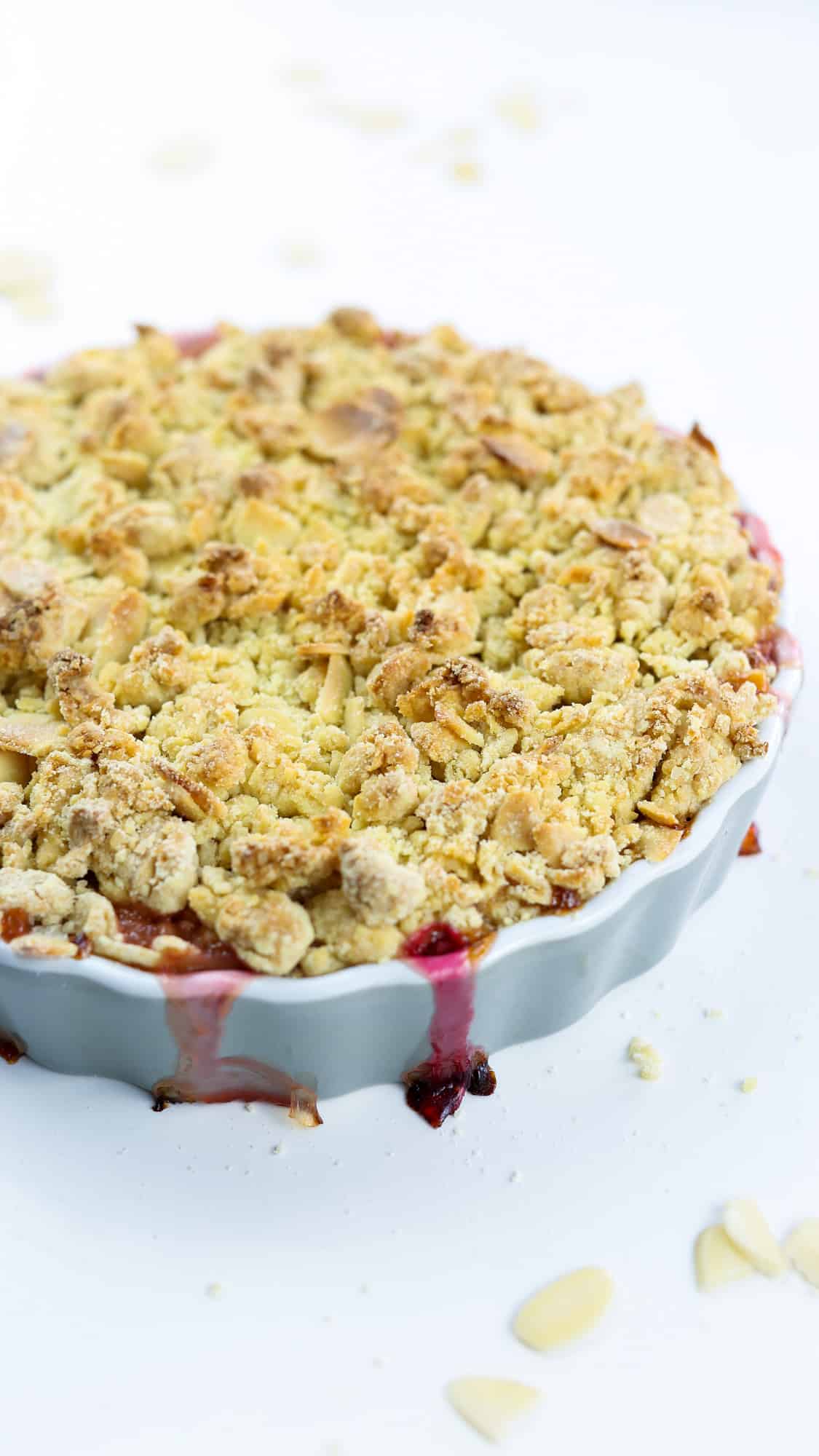 vegan Amaretto & Rhubarb crumble on a grey, round ceramic dish on a white surface with almond shavings