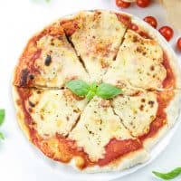 whole vegan pizza margherita cut in 6 slices with fresh basil leaves in the centre on a white background decorated with ripe cherry tomatoes and basil leaves