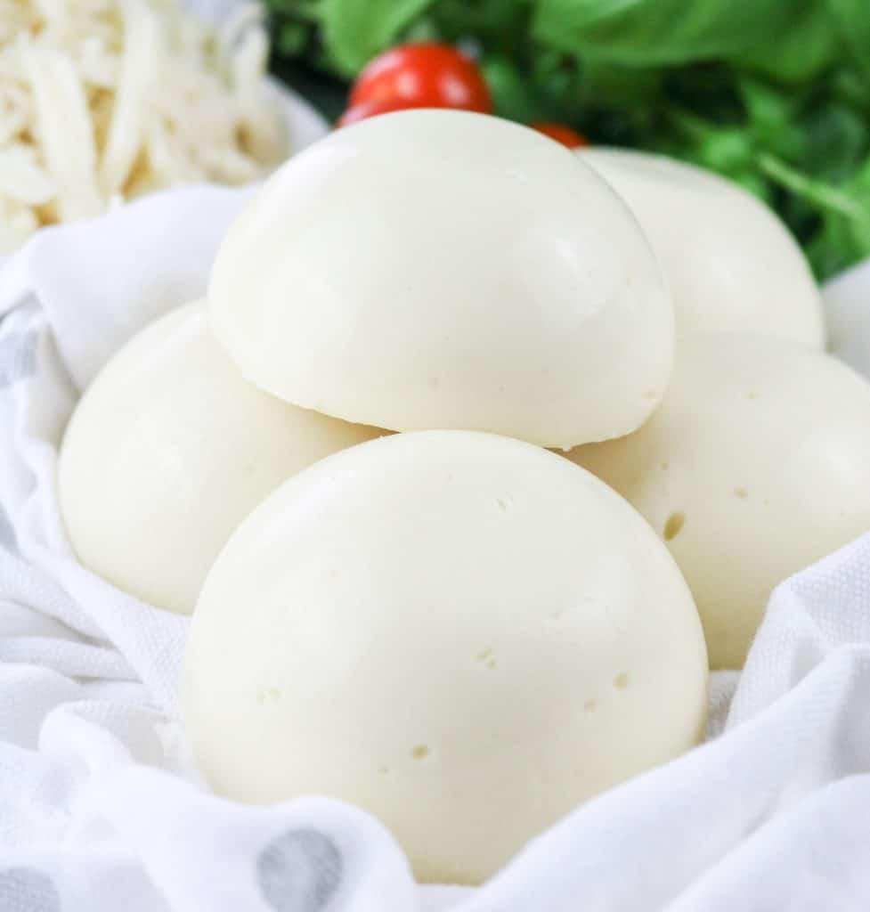 Vegan Mozzarella semi spheres on a white cloth with grey polka dots on a background of grated mozzarella shavings, cherry tomatoes and fresh basil