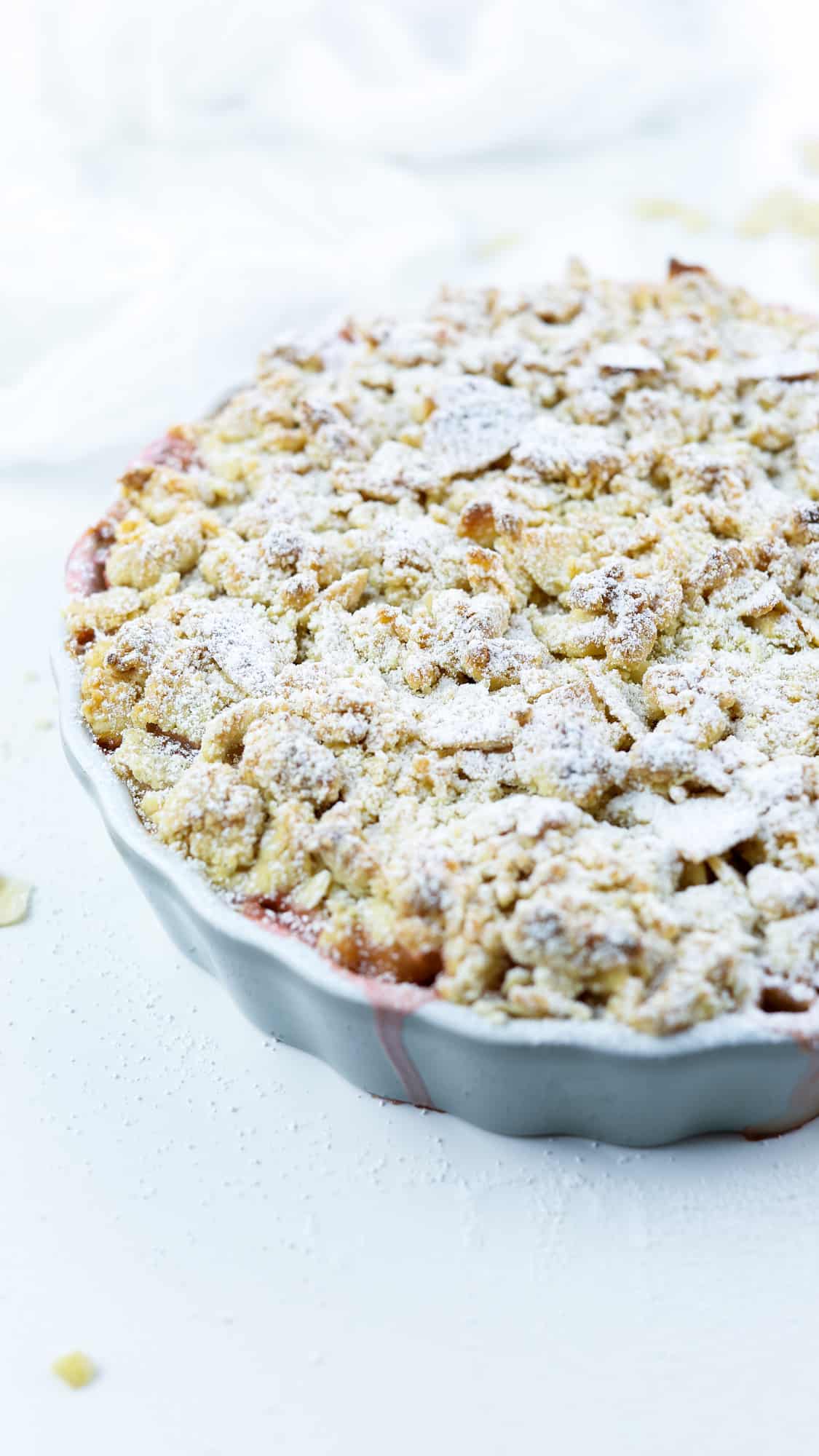 vegan Amaretto & Rhubarb crumble on a grey, round ceramic dish on a white surface with almond shavings with a light icing sugar layer on top