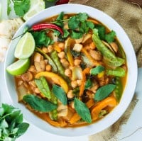 White ceramic bowl filled with rich vegan thai red curry with chickpeas, peppers, mangetout, thai basil, coriander leaves and lime wedges