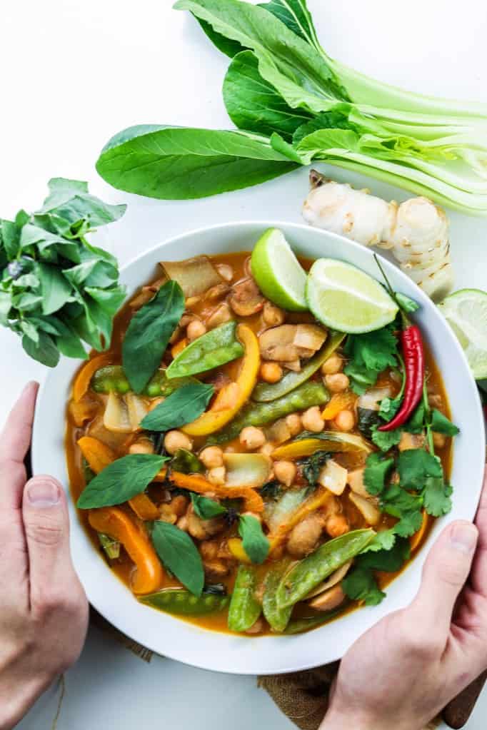 White ceramic bowl hold by both hands filled with rich vegan thai red curry with chickpeas, peppers, mangetout, thai basil, coriander leaves and lime wedges