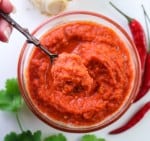 Vegan Thai Red Curry Paste in a glass bowl on a white table with chillies, coriander leaves and galangal root focus on a copper spoon hold with left hand from the top left corner