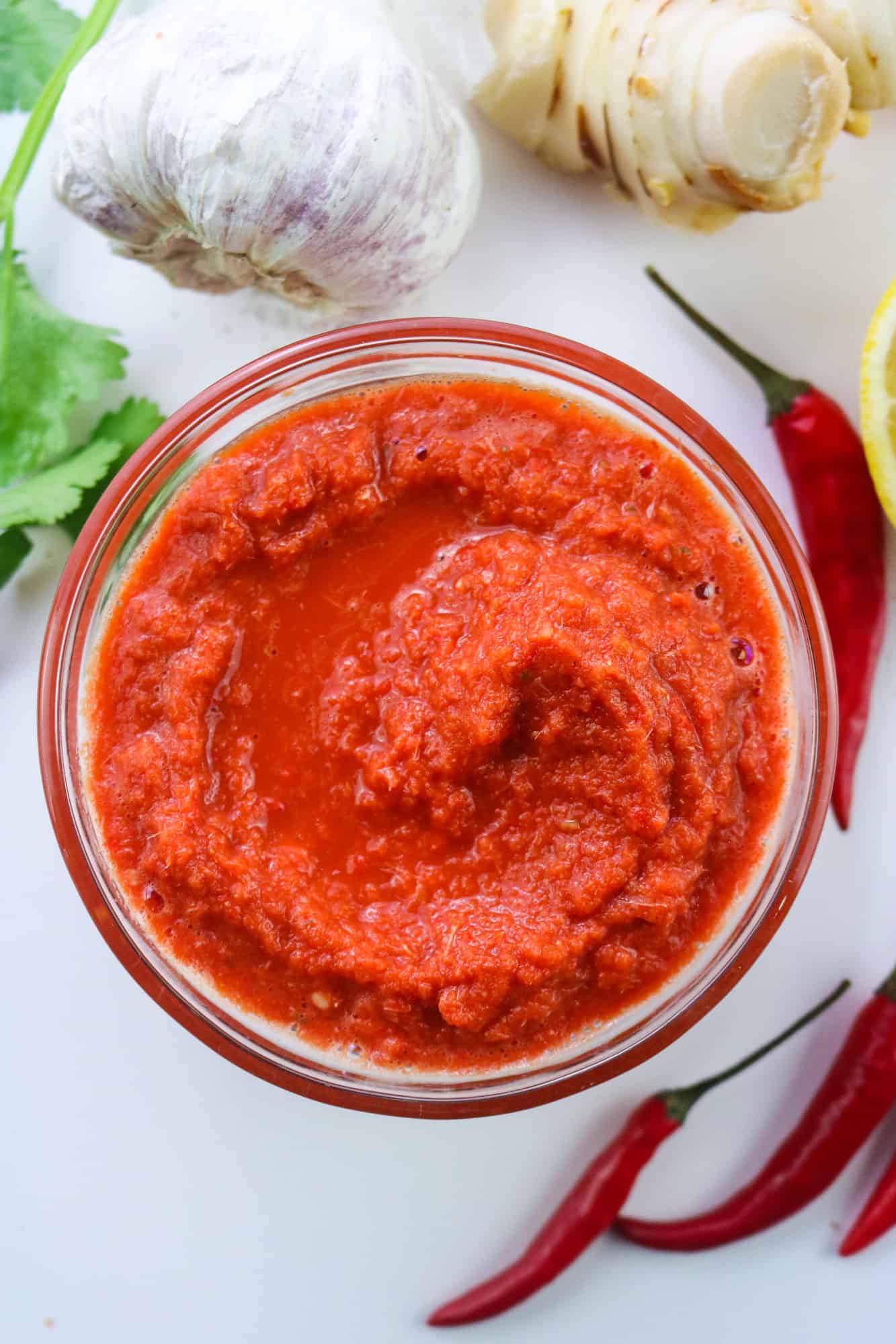 Vegan Thai Red Curry Paste in a glass bowl on a white table with chillies, coriander leaves and galangal root, garlic bulb and lemon wedge