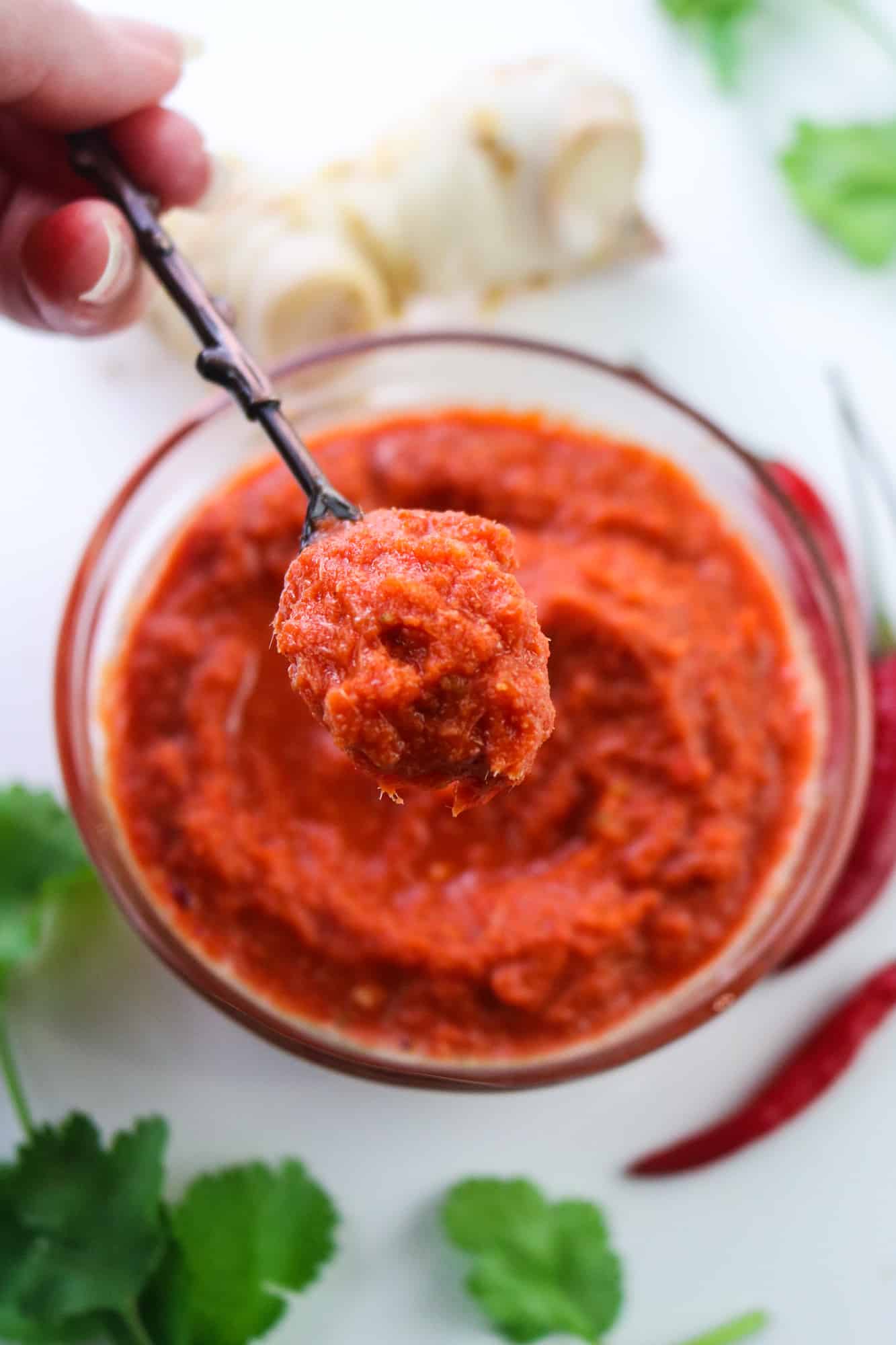 Vegan Thai Red Curry Paste in a glass bowl on a white table with chillies, coriander leaves and galangal root focus on a copper spoon hold with left hand from the top left corner
