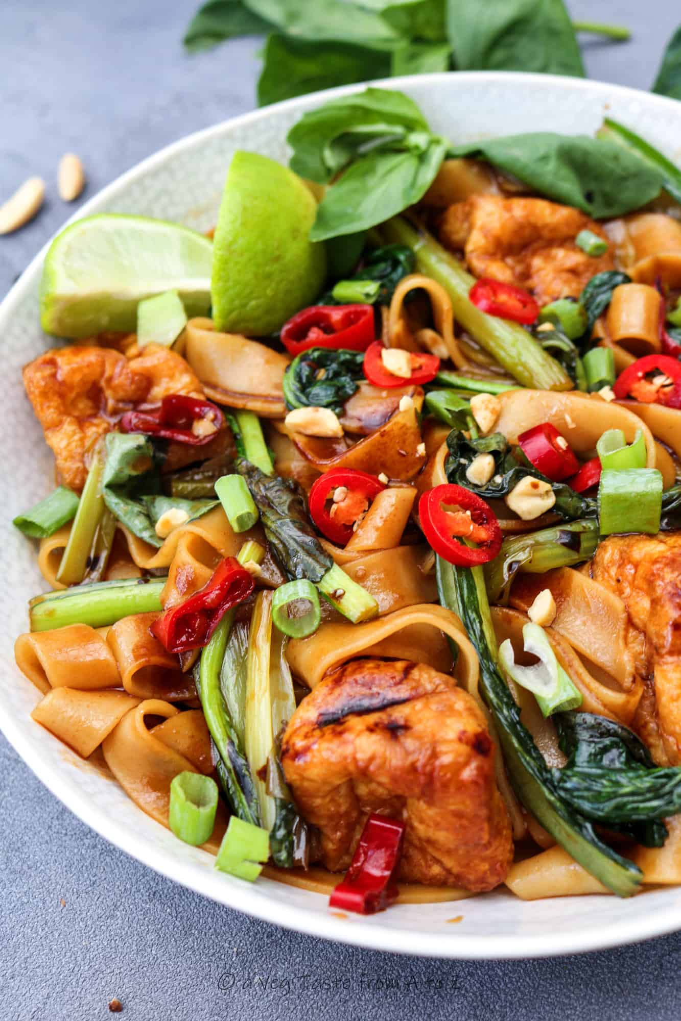 rice noodles, greens and puffed tofu in white ceramic bowl on brown cloth filled with rice noodles, greens and puffed tofu with chopsticks in the plate on white table decorated with half pak choi and thai basil close up