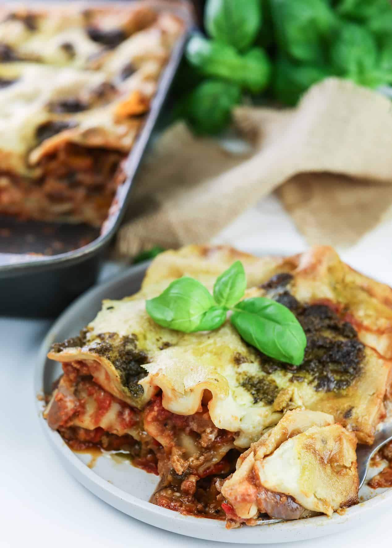 slice of creamy veggie lasagna with pesto and mozzarella sauce topped with basil leaves with entire tray and basil plant on the background