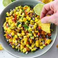 colourful mango salsa in a grey contrasting plate with hand holding a tortilla chip ready to dig in