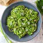 wild garlic pesto spaghetti rolled into nests served on a grey plate and topped with pine nuts and wild garlic flowers
