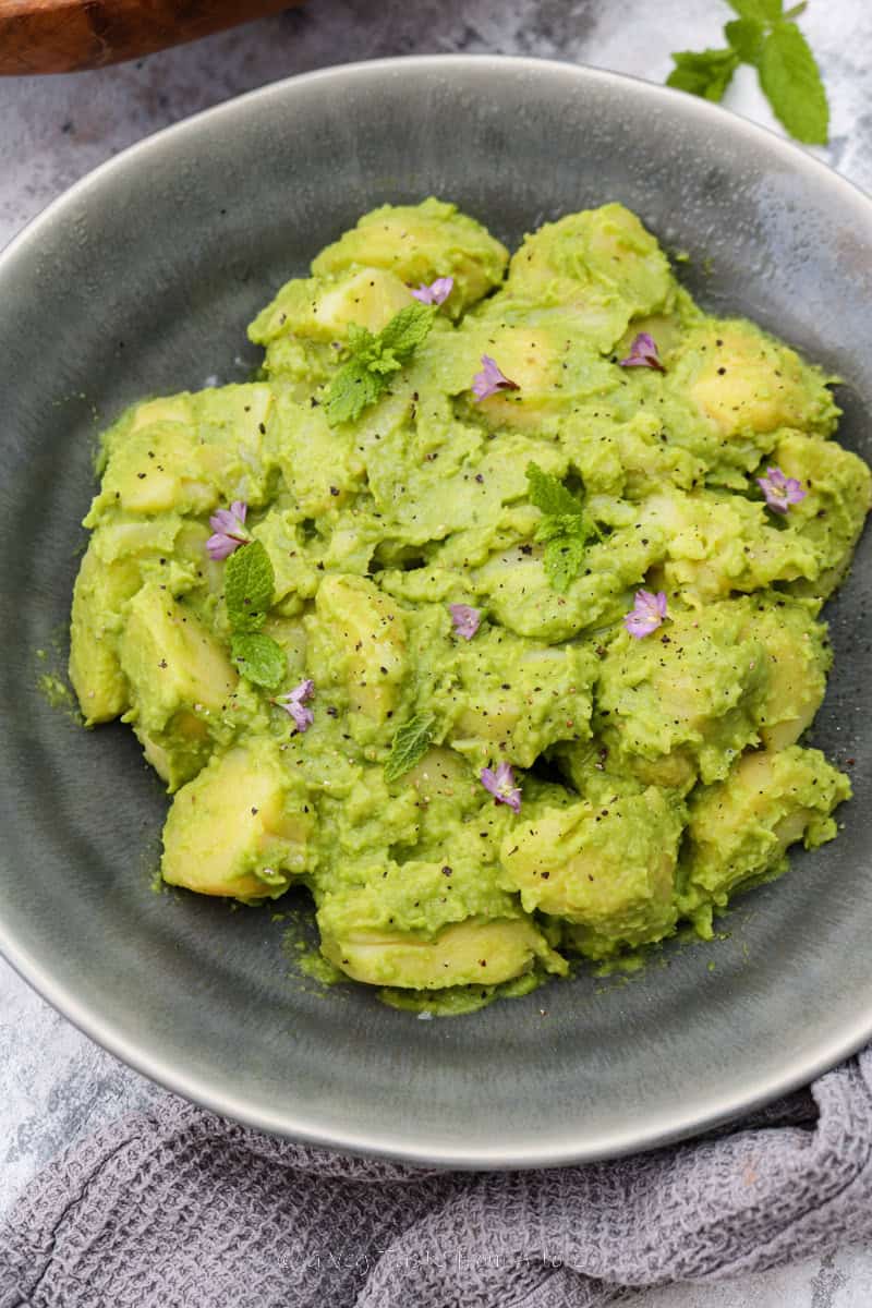 close up to half plate of boiled potatoes chunks coated in pea and mint sauce in a grey dish over a grey background garnished with fresh mint leaves and purple flowers