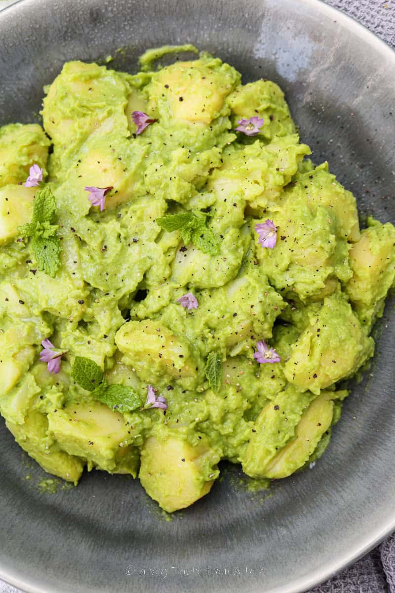 close up to half plate of boiled potatoes chunks coated in pea and mint sauce in a grey dish over a grey background garnished with fresh mint leaves and purple flowers