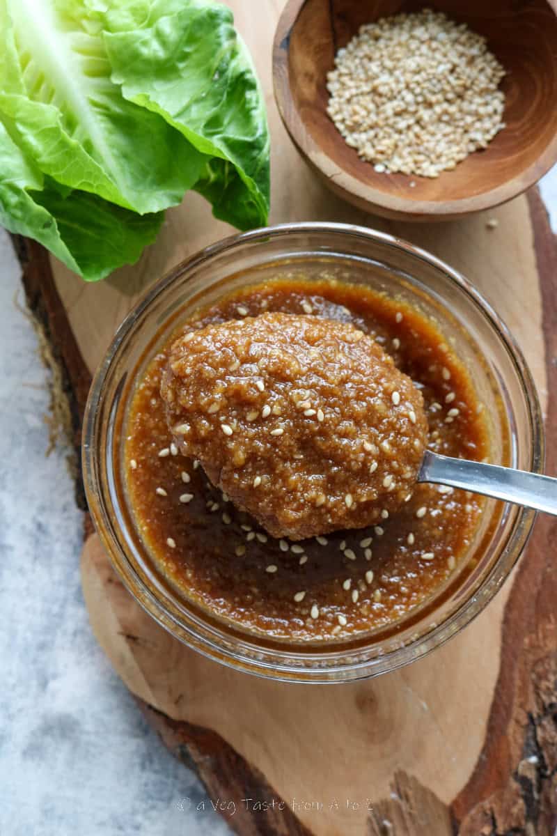 sesame and miso dressing in a glass bowl with a spoon over a wooden board with sesame seeds and salad props