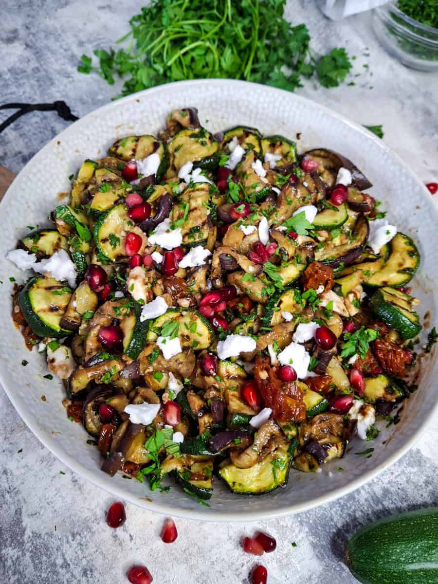 vibrant and healthy Mediterranean grilled veggie salad, loaded with charred aubergines, courgettes, vegan feta, sundried tomatoes, and pomegranate seeds on a serving plate